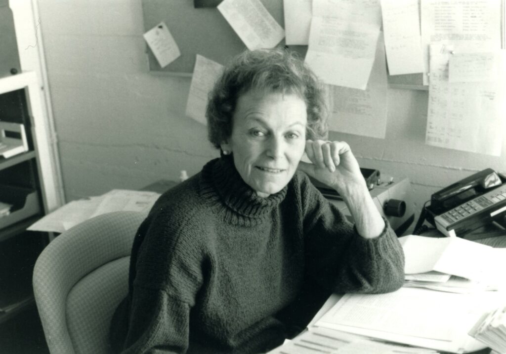 Ploughshares founder Sally Lilienthal smiles from her desk, papers pinned to the wall behind her